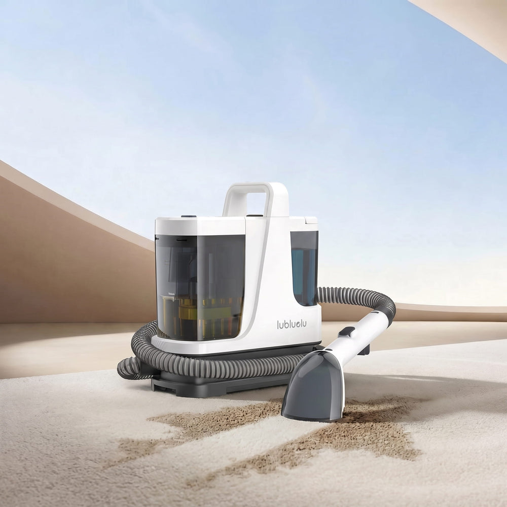 A white and gray LuluBlue carpet cleaner with a flexible hose and handheld attachment, shown cleaning a beige carpet in a stylized desert-like setting with sand dunes and a blue sky background.